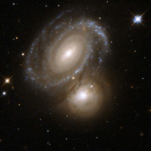 Spiral galaxy above and slightly left of center, blue-white arms made of of stars, and streaks of brown gas. To its lower right, just below central, an orange-white glowing sphere. Bright foreground star in the upper-right.
